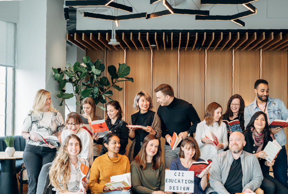 Group of CZI Education Design team members pose indoors, smiling and looking through books (Summit Learning Platform, Annual Letter).