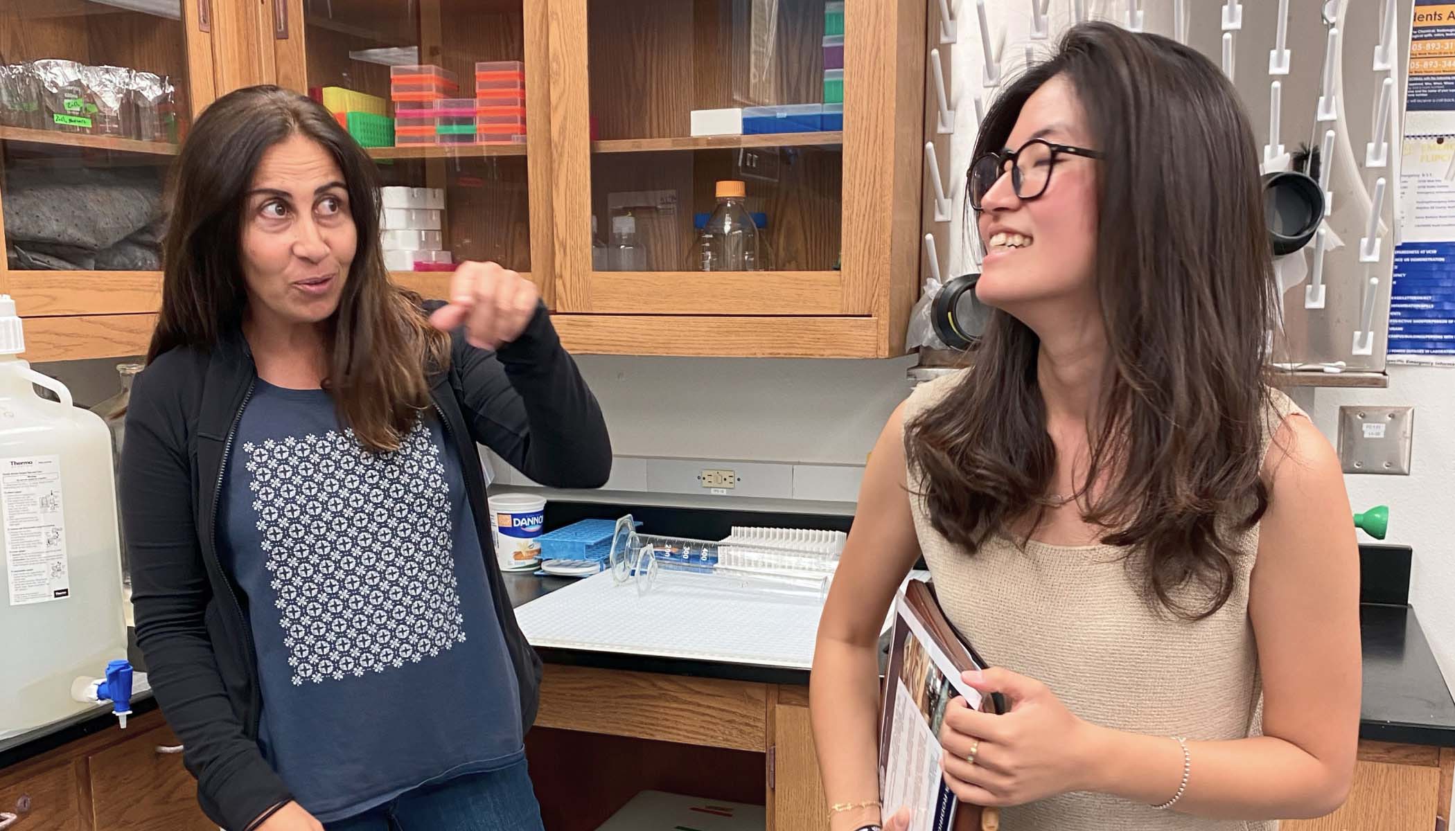 Two women in conversation in a lab. The woman on the left wears a black jacket. The woman on the right wears glasses and a beige top.
