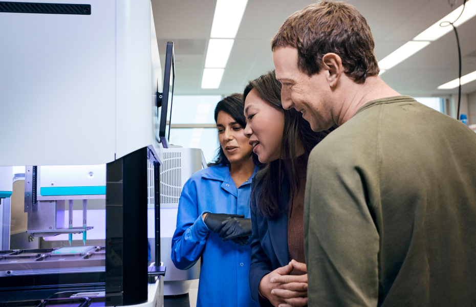 Inside a lab with modern instruments and monitors visible, a scientist in a blue lab coat gestures toward a large piece of equipment while two visitors lean in to observe closely.