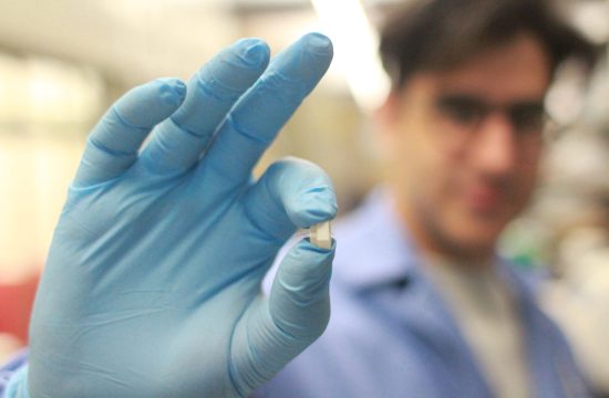 Gloved hand holding a small medical sensor in a lab, researcher blurred in the background.